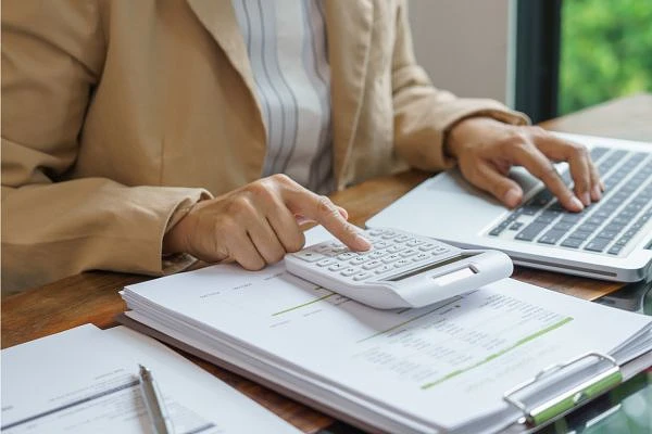 A person is using a calculator while working on a laptop and examining documents on a desk.