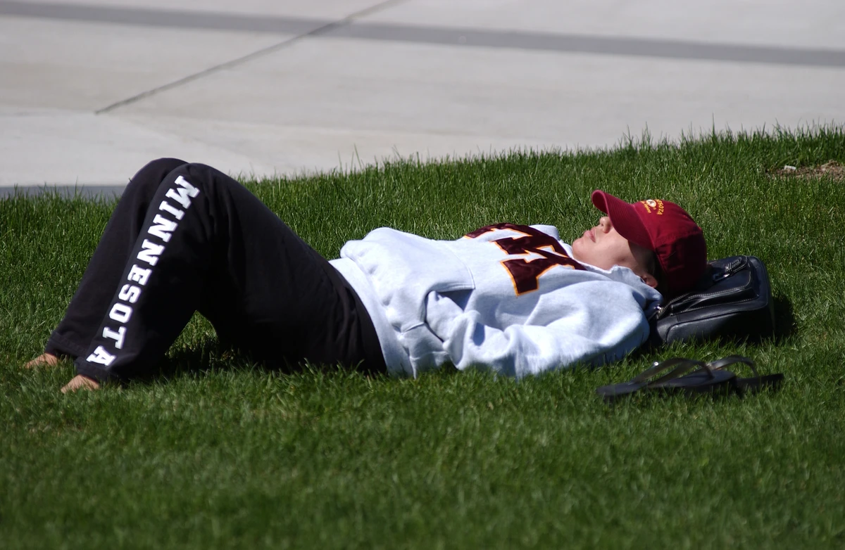 A student wearing "Minnesota" sweatpants and a marron cap is lying on grass, relaxing in the sun with their eyes closed.